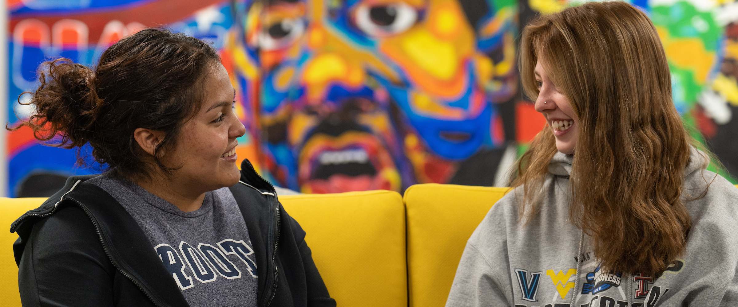 Two students gathered and chatting with a vibrant painting in the background.