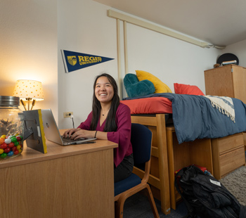 Student at her desk in her dorm room, smiling while working on her laptop
