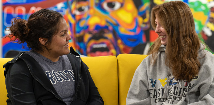 Two students sit down side by side on a couch and look at each other while smiling