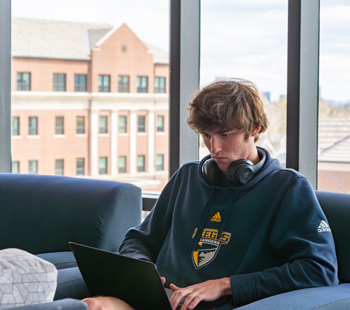 Student sitting and focused on their laptop, headphones around the neck, in one of the common spaces in Desmet Hall