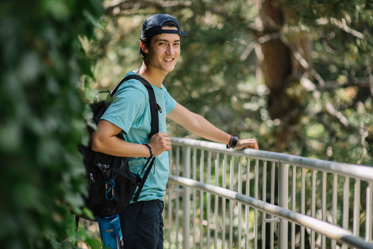 A student with a backpack and a water bottle smiles while leaning on a railing in a green outdoor setting