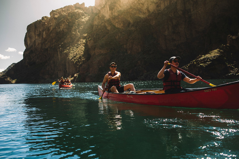 Two students paddling a red canoe on a scenic river canyon with dramatic sunlit rock cliffs in the background, with additional kayakers visible in the distance.