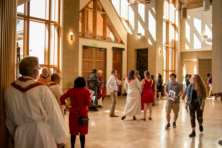 Students and faculty mingling in the sunlit atrium of a modern chapel or religious building, with clergy in vestments and attendees in formal attire conversing after a service or ceremony.
