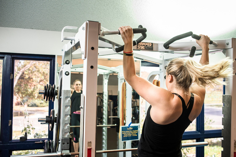 A woman doing pull-ups on a Hammer Strength cable machine in a campus fitness center, with her reflection visible in a mirror in the background.