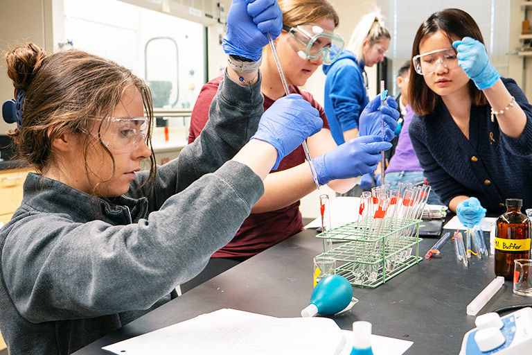 undergraduate students handle use pipettes to fill vials while wearing eye protection and hands during a chemistry lab in a Regis classroom