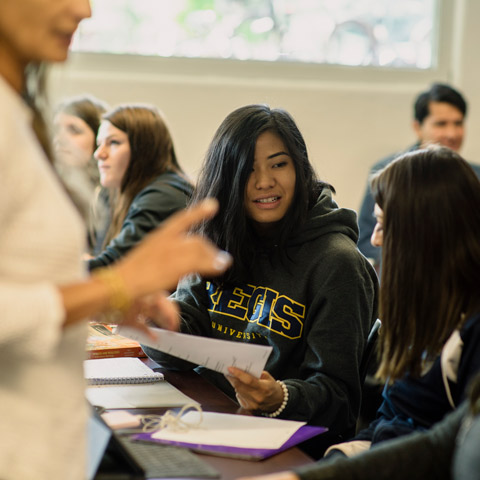 Students collaborating in a classroom at Regis University, reviewing course work together while an instructor leads the discussion.