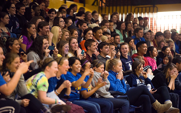 a crowd of students sit in gym bleachers laughing, smiling and taking photos with their phones