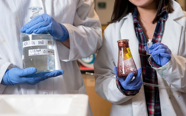 2 people wearing lab coats hold beakers containing fluids