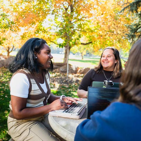 Regis University students collaborating with laptops at an outdoor table on campus.