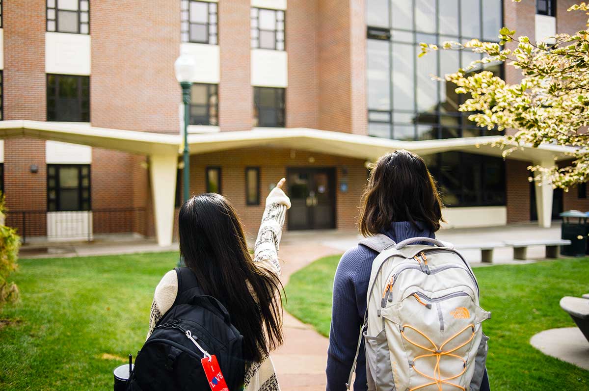 two students wearing backpacks walk on the sidewalk on the Northwest Denver campus