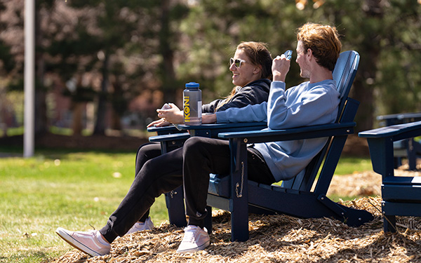two students chat and recline in the sun in lawn chairs on the Quad