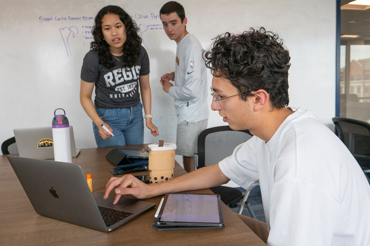 Three students work together on a project in a classroom on the Regis campus, typing on a laptop and writing on a white board