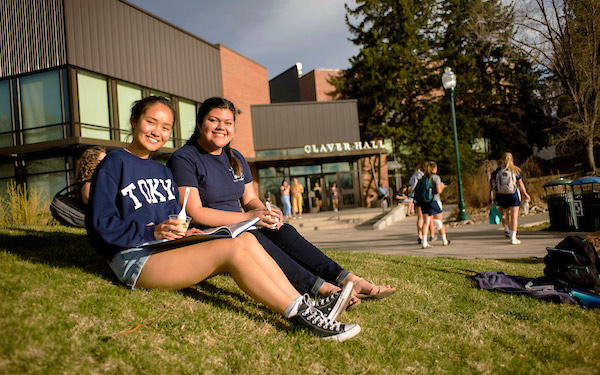two students sit in the grass with open books and snacks outside Claver Hall