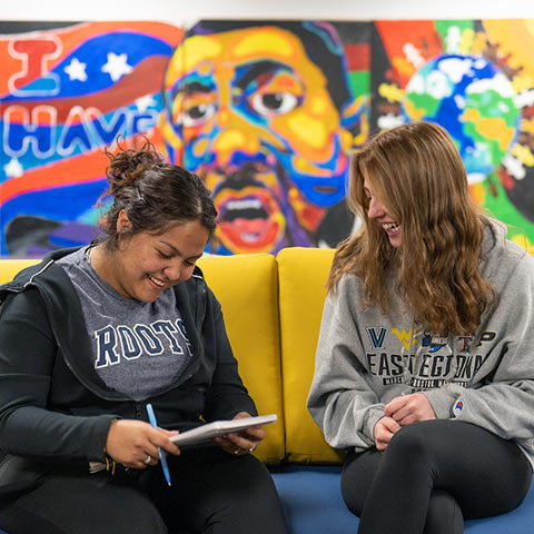 Two young women write in a notebook and sit on a sofa in front of a mural of MLK that reads I Have a Dream