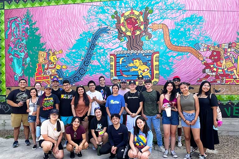 large group of students smile in front of colorful mural