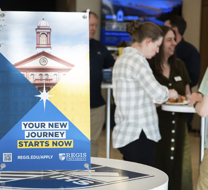 Banner on left of photo: Your New Journey Starts Now, REGIS.EDU/APPLY. Multiple people are in the background of the right side socializing