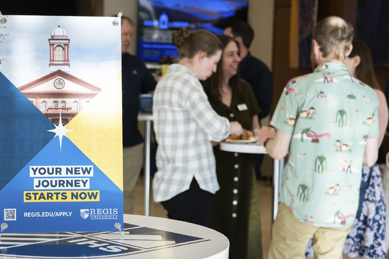 Regis banner on the left: Your New Journey Starts Now, Regis.edu/apply. Group of adults on the right interact around a round standing table