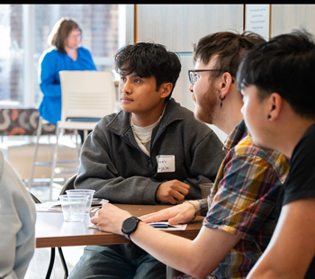 Three students sit at a table listening to someone on the left of the image