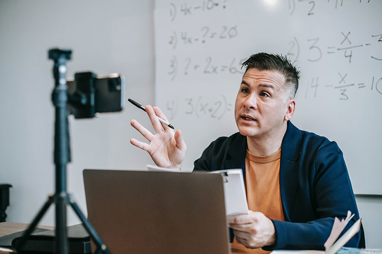 teacher sits at a desk in front of a whiteboard while talking to a camera and using a laptop