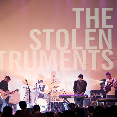 Four members of The Stolen Instrument play their instruments, including guitars, drums and keyboard, on stage in front of an audience.