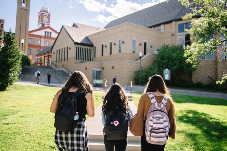 Three students with backpacks walk toward a sunlit campus building with a bell tower
