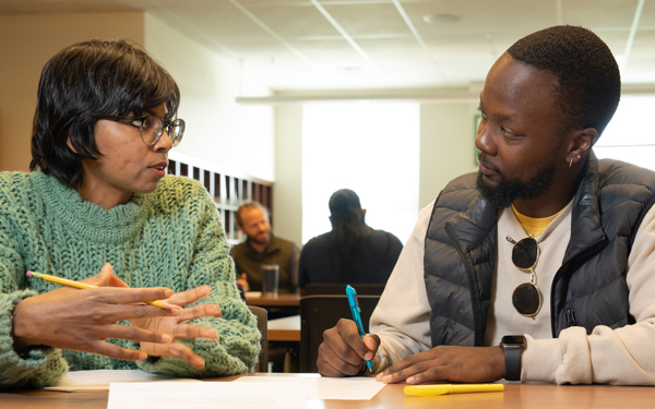 Two students sit at a table in a study area, discussing notes. One gestures while speaking, and the other listens, holding a pen.