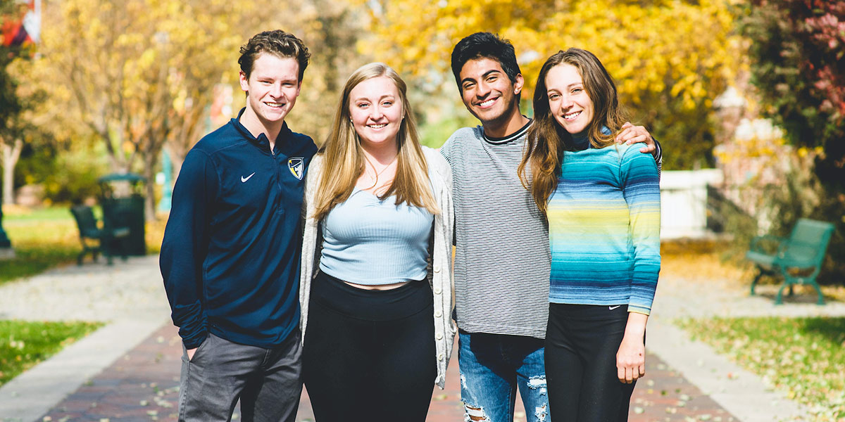 Four smiling students stand arm-in-arm on a tree-lined campus path during fall, surrounded by golden autumn leaves.
