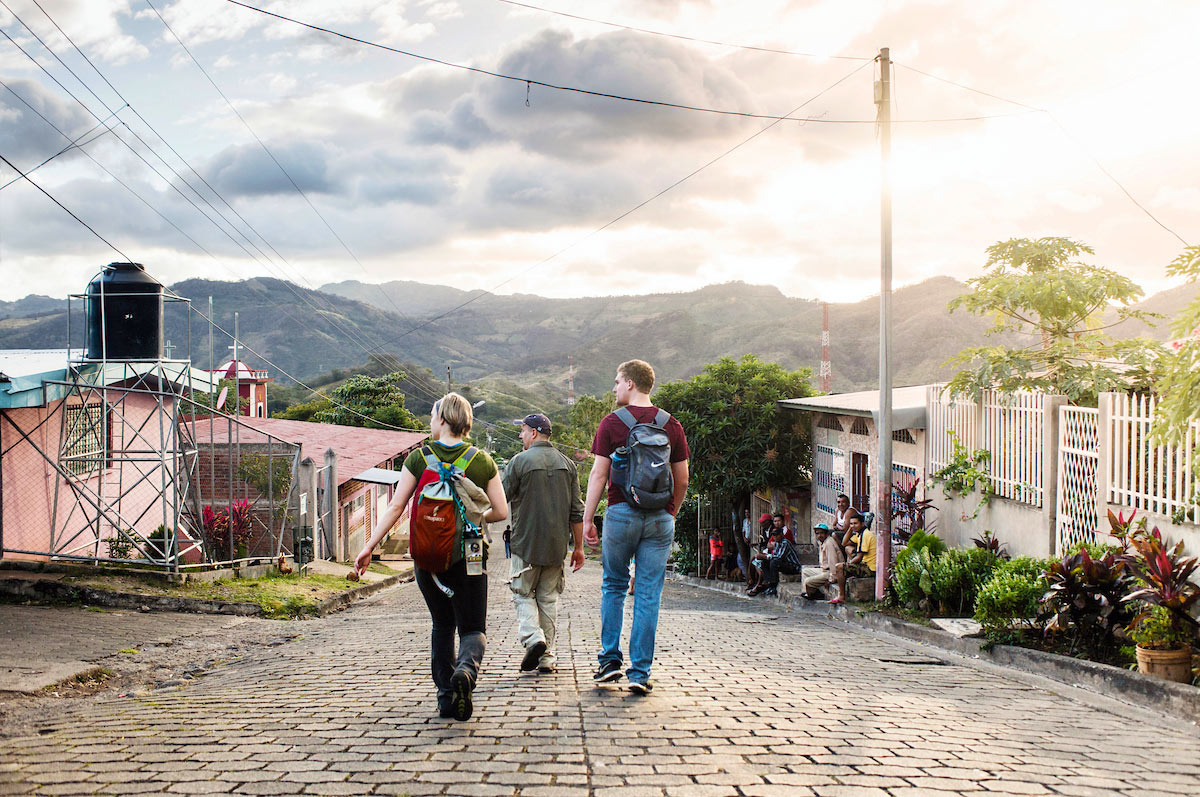 students wearing backpacks follow their chaperone as they walk down a cobblestone street in a town with mountains stretching into the distance in Nicaragua