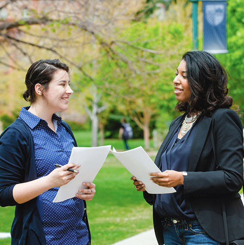 two women hold papers while standing outdoors and talking to each other