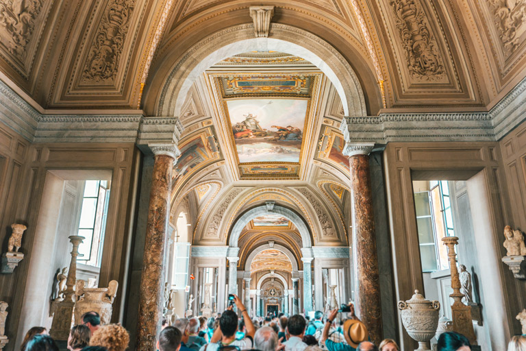 Crowd of people walking through a museum in Vatican City, featuring arched ceilings, marble columns, classical statues, and decorative ceiling paintings