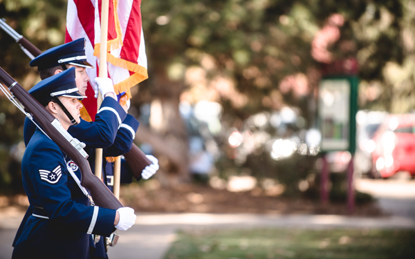 Three uniformed members of a color guard march in formation, carrying rifles and the American flag during an outdoor ceremony on campus.
