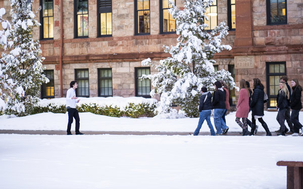 Snow covers the grass and lays on the branches of trees. A Regis tour guide walks in front of a group of people as they walk on a paved sidewalk