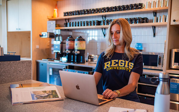 woman wearing a Regis t-shirt works on a laptop in a coffee shop