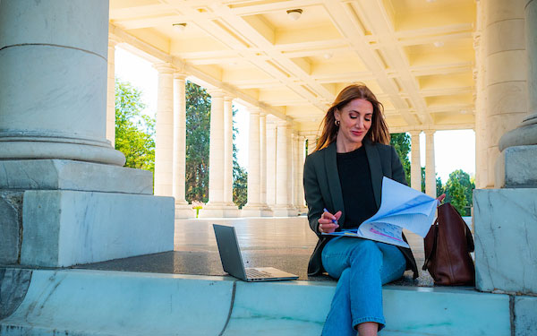 young professional woman writes on a notepad with her open laptop beside her while sitting in an urban park