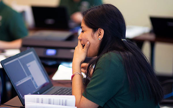 a woman smiles while reading from a textbook in front of her laptop in a classroom