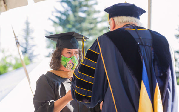 Graduate in cap, gown, and colorful face mask receives diploma from university official in academic regalia during Regis University's 2020 commencement ceremony.