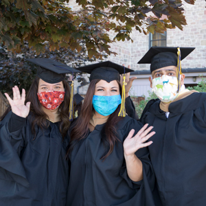 Regis University students in graduation caps and gowns celebrating commencement during the COVID-19 pandemic, wearing colorful face masks and waving.
