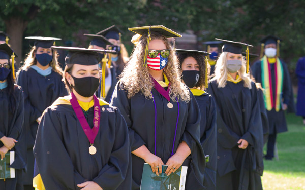 Graduates wearing caps, gowns, medals, and face masks—including one with an American flag design—stand outdoors during Regis University's 2020 commencement ceremony.
