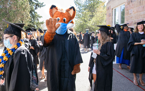 "Graduates in caps, gowns, and face masks gather outdoors during Regis University's 2021 commencement ceremony, with Regi the Ranger fox mascot also in a gown and mask, waving at the camera.
