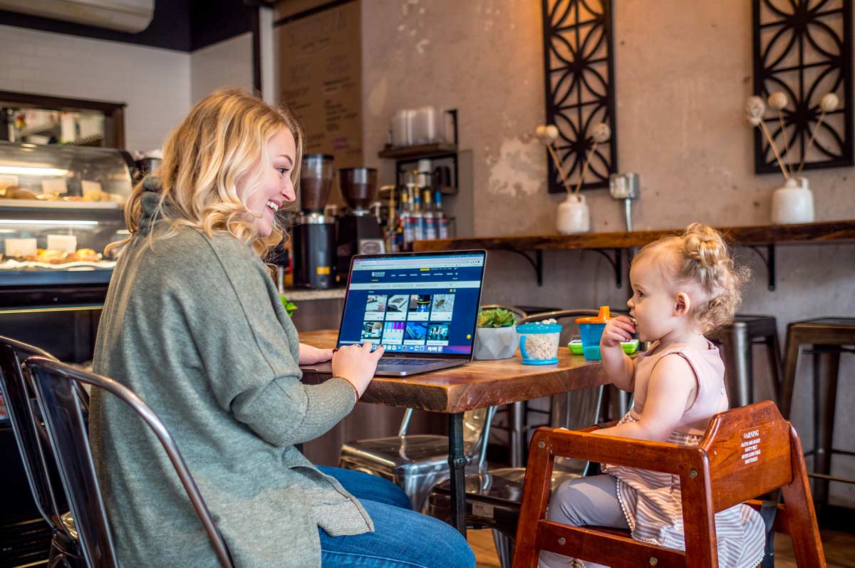 Adult learner on the left has their laptop open on the Regis University website and is sitting next to their child on the right. In a small coffee shop