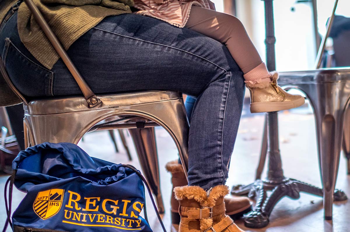 An adult student with a child sitting on her lap studies at a café, with a Regis University tote bag at their feet.
