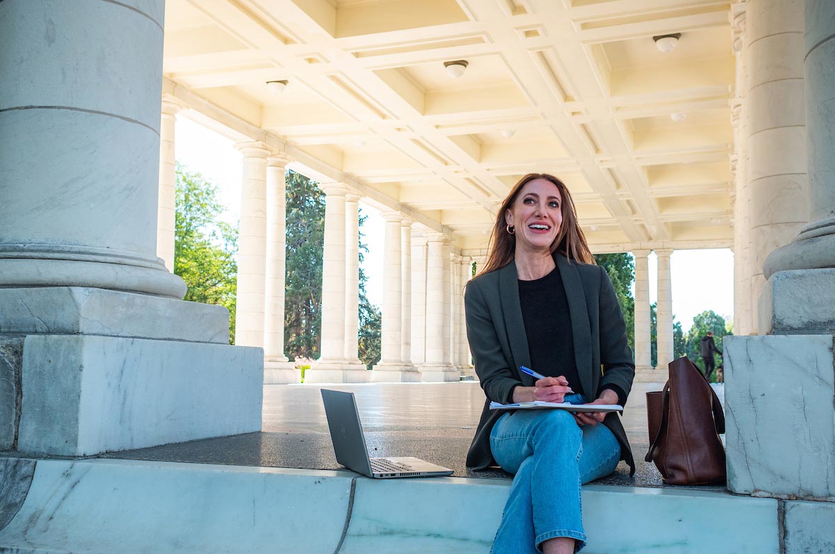 A woman sits under a marble archway in a park with a notebook and pen in hand and laptop next to her, studying for her class at Regis University in Denver, Colorado.