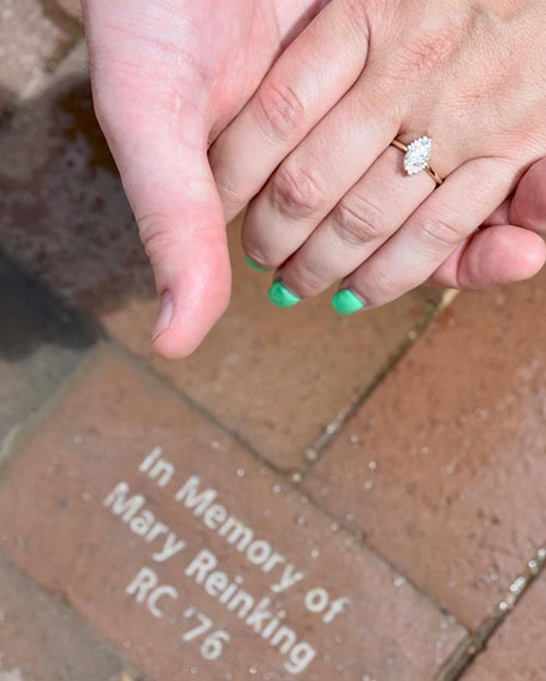 Anne and Bobby hold hands, Anne's hand sports a diamond ring and a memorial brick in the background reads "In Memory of Mary Reinking RC '76"