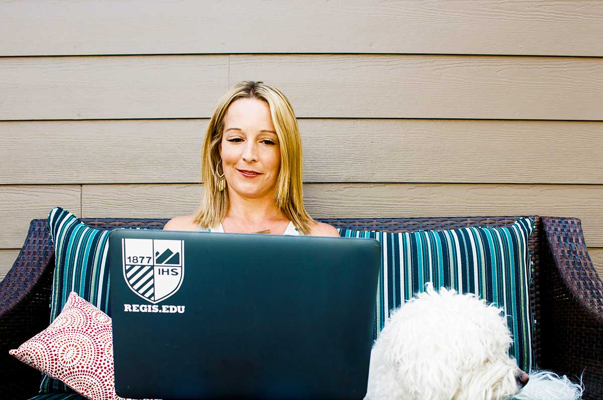 adult woman sits on a sofa on her patio working on a laptop with her dog by her side