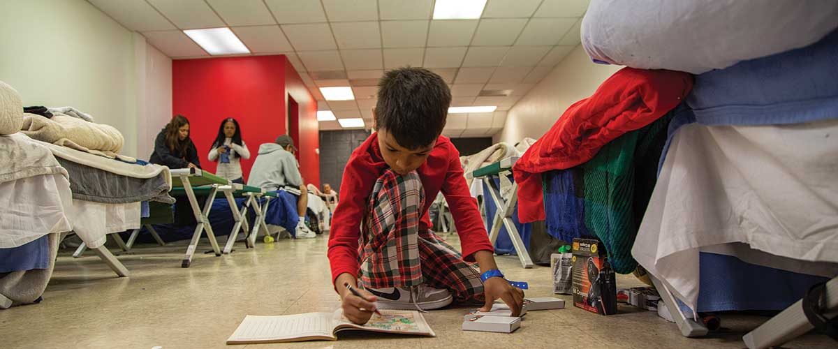 A small boy sits on the floor enjoying crayons and a coloring book among cots at the Regis Welcome Center.