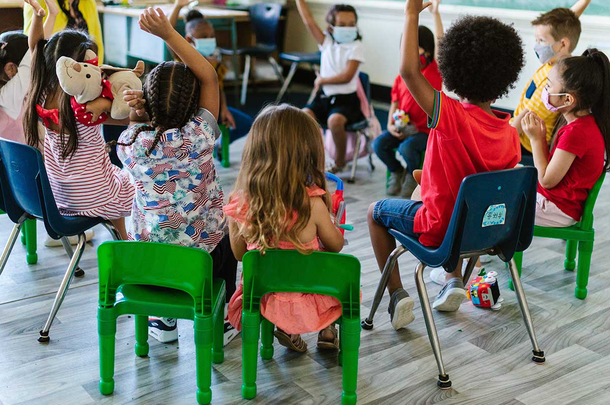 children sit in chairs in a circle with their hands raised in a colorful classroom