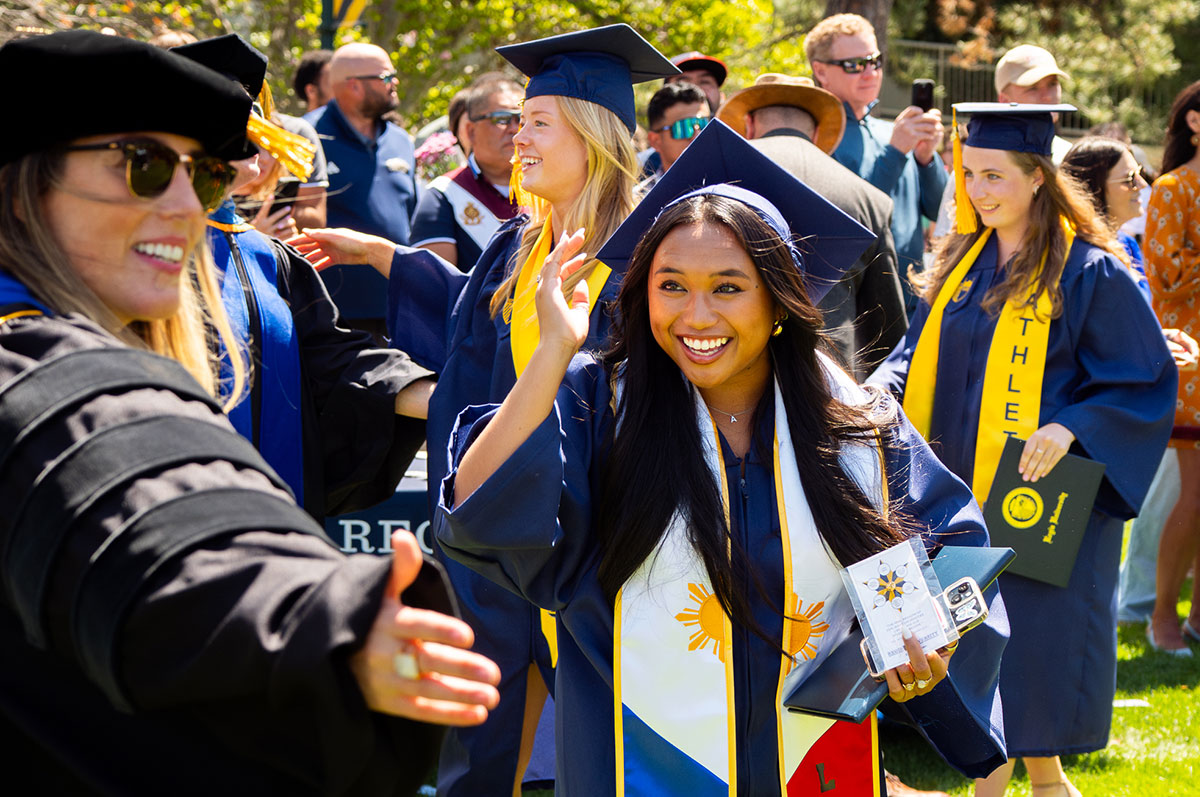 wearing her graduation cap and gown and smiling from ear to ear, a young woman clutches her diploma and process from the stage at her commencement ceremony on the Regis campus