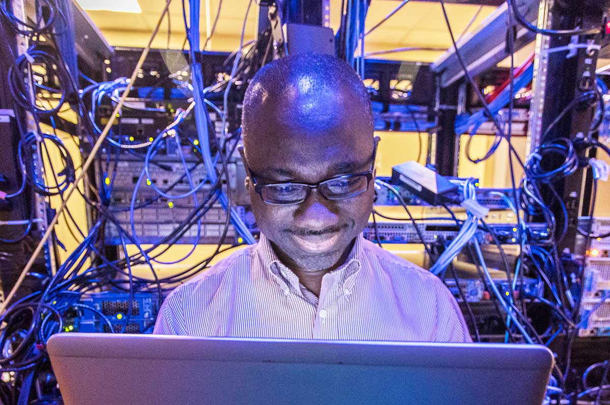 a man smiles while working on a laptop in a server room