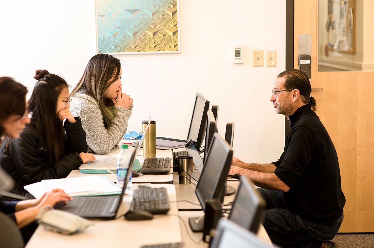 students sit at computers while a professor crouches down and coaches them