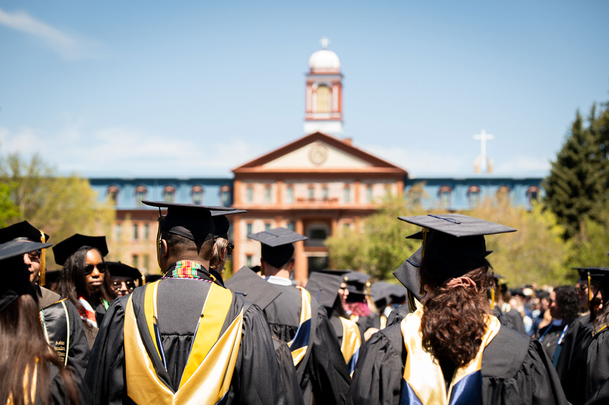 A photo of graduates in caps and gowns, seen from behind, gathered outdoors in front of Regis University Main Hall, on a sunny day.